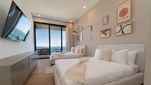 Une chambre d’hôtel moderne dotée d’un grand lit, d’un plus petit lit, d’un téléviseur fixé au mur et offrant une vue sur la mer grâce à de grandes fenêtres.