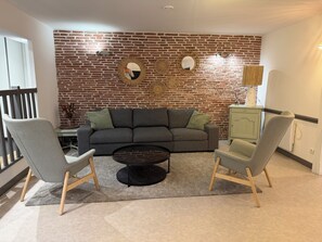 A living room with a brick wall, a grey sofa, two armchairs, a coffee table, and a cabinet. - Le Petit Sancerrois – a loft in the heart of historic Sancerre (Sancerre)