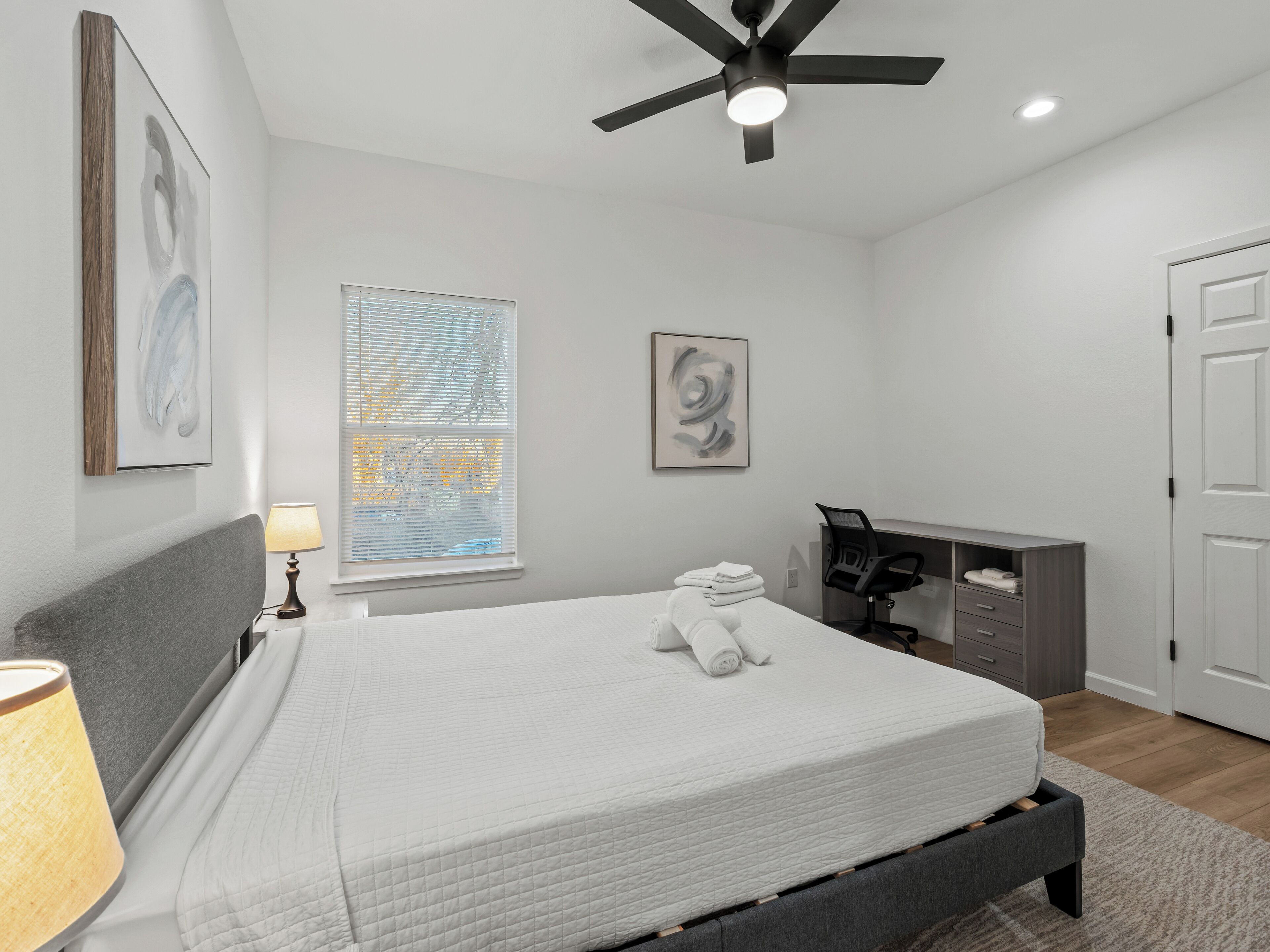 Une chambre à coucher comprenant un lit, un bureau, une chaise, un ventilateur de plafond et des œuvres d’art accrochées aux murs.