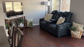 Ein Wohnzimmer mit dunklem Holzboden, einem schwarzen Ledersofa, einem Flachbildfernseher auf einem Holzständer und einem Essbereich mit Holztischen und einer Tischplatte.