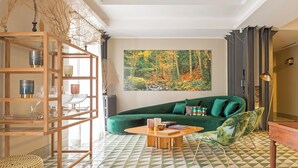 Een woonkamer met een groene bank, een houten boekenkast en een groot schilderij met een boslandschap op de muur.