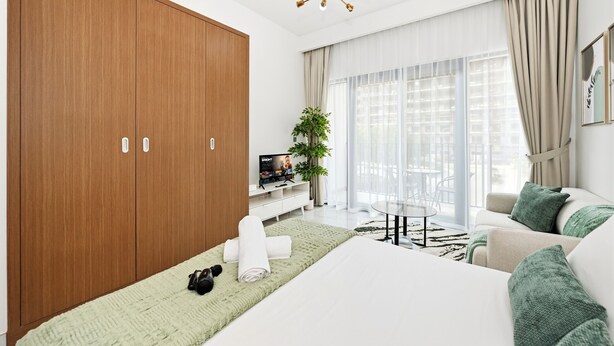 Ein modernes Hotelzimmer mit großem Kleiderschrank, einem Bett mit grüner Decke, einer Couch, einem Fernseher und einer Pflanze.