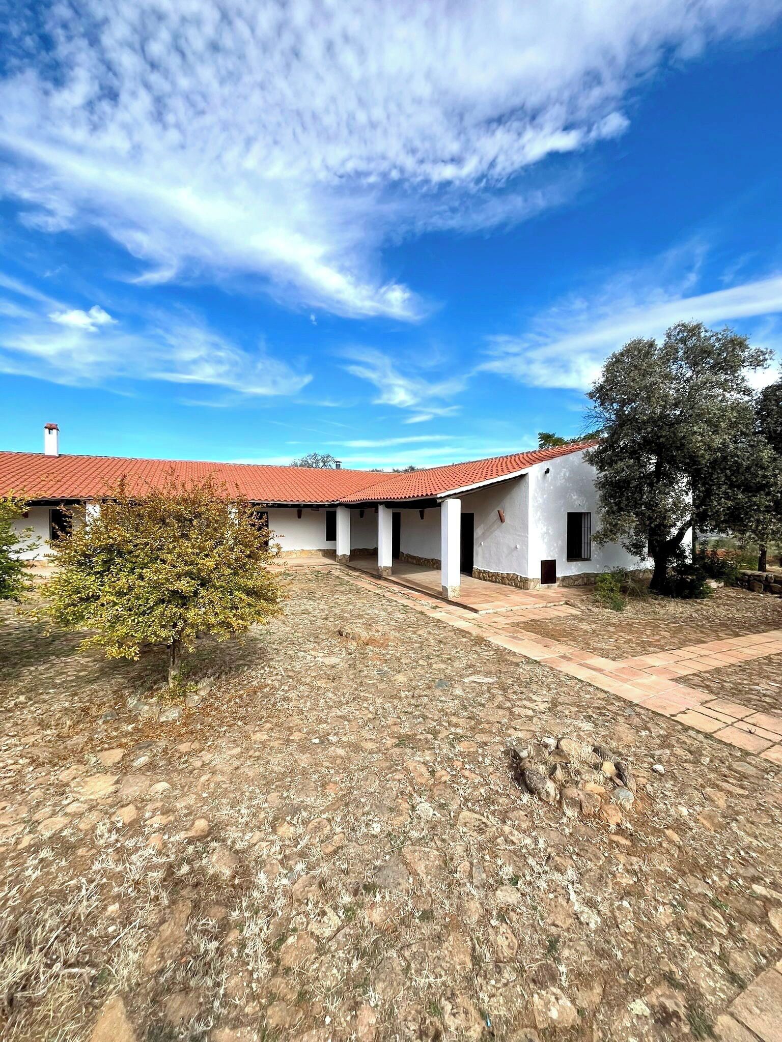 Un edificio con techo de tejas rojas, paredes blancas y un patio con pavimento de piedra y vegetación escasa.