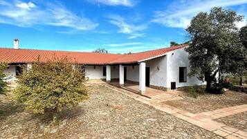 Un edificio con techo de tejas rojas, paredes blancas y un patio con pavimento de piedra y vegetación escasa.