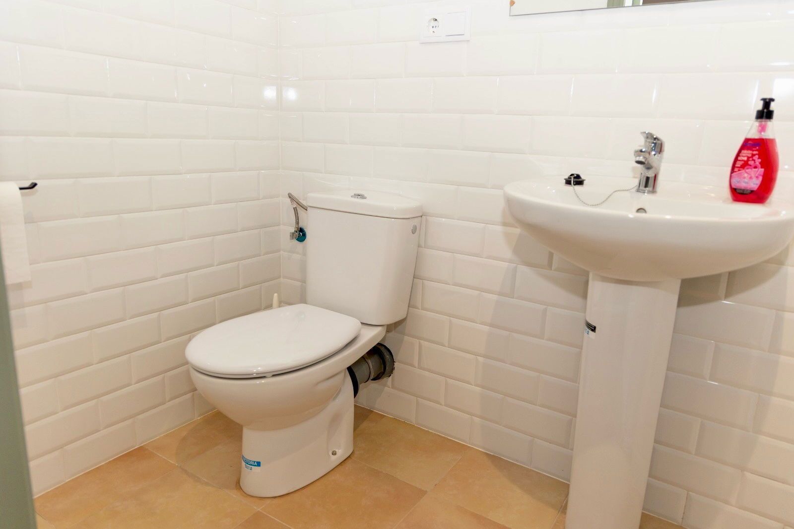 A small bathroom with white tiles, a white toilet, a sink, and a red bottle of soap.