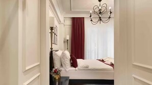 Een slaapkamer met een bed, een nachtkastje, een vaas met bloemen, een kroonluchter en een raam met gordijnen.