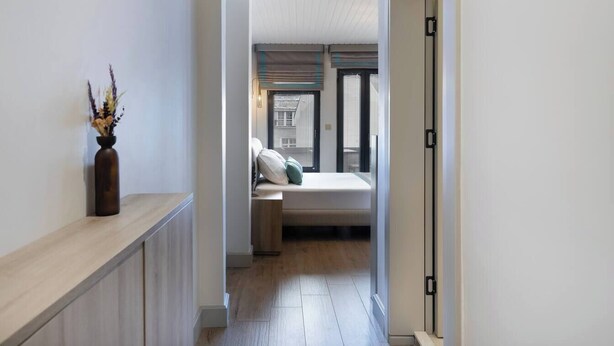 A narrow hallway with a wooden floor leading to a bedroom with a bed, a nightstand, and a window with blinds. - The One Taksim 105 Room, Comfort & Privacy (Beyoglu)