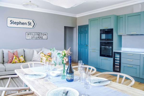Contemporary living room with a plush beige sofa and patterned cushions, accented by quirky street-sign décor. Warm lighting and cloud-like fixtures add charm, creating a cozy, stylish space that flows into the kitchen and dining area.