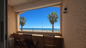 A room with a large window overlooking a beach, two wooden chairs, and a small trash bin. - Front-line studio cabin (Fleury)