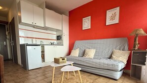 A compact living space with a kitchenette, a grey sofa, a small round table, and a red accent wall. - Front-line studio cabin (Fleury)