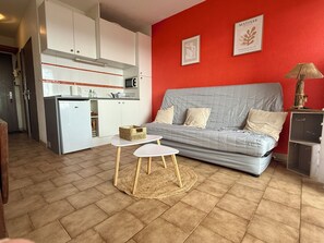 A compact living space with a kitchenette, a grey sofa, a small round table, and a red accent wall. - Front-line studio cabin (Fleury)