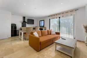 A modern living room with a sofa, a coffee table, and a kitchen area with a dining table and chairs. - La Volière du Pesquier – Holiday Home in Provençal Pine Forest with Pool and Garden (Pélissanne)
