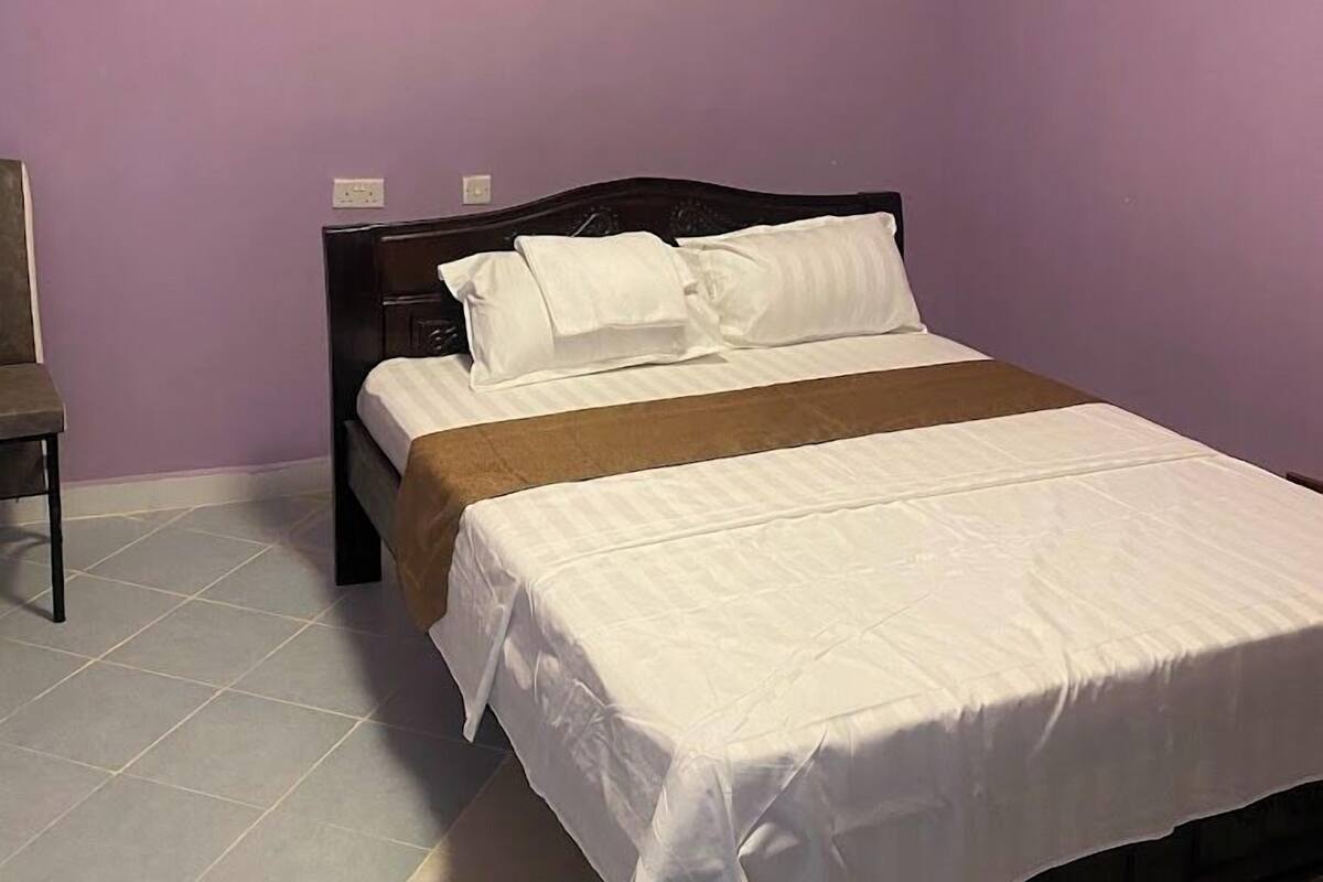Una cama individual con ropa de cama blanca en una habitación con paredes moradas y piso de baldosas.