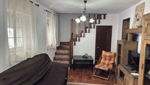 Un salon avec un canapé marron, un meuble TV en bois, un fauteuil à motifs, une petite table avec une tasse et un escalier surmonté d’un lustre.