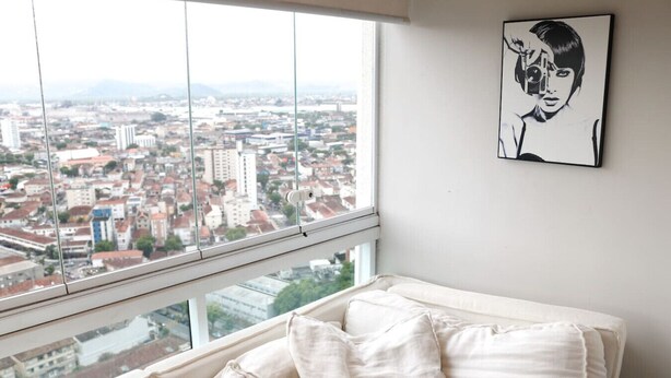 A bedroom with a large window overlooking a cityscape, a bed with white bedding, and a black and white portrait on the wall. - Cobertura de Alto Padrão • Cyrela • Boqueirão (Santos)