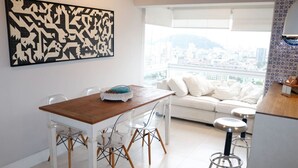 A modern kitchen with a wooden table and chairs, a white sofa, and a city view through large windows. - Cobertura de Alto Padrão • Cyrela • Boqueirão (Santos)