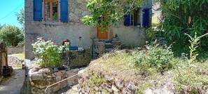 Ein rustikales Steinhaus mit blauen FensterlÀden, einer Steintreppe und einem Garten mit rosa Blumen.
