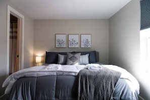 Ein ordentlich bezogenes Bett mit grau-weißer Decke, einem dunklen Kopfteil und drei gerahmten floralen Kunstwerken an der Wand.