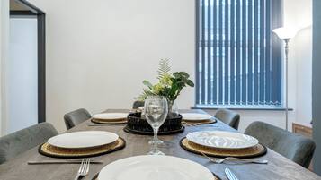 A dining table set with plates, cutlery, and a glass, with a plant centerpiece and a window with blinds in the background.