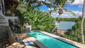 A rectangular pool with a stone border, surrounded by lush greenery and palm trees, overlooking a calm body of water. - A Casa do Lago: comfort, nature and unforgettable moments on the shore of the water (Romana)