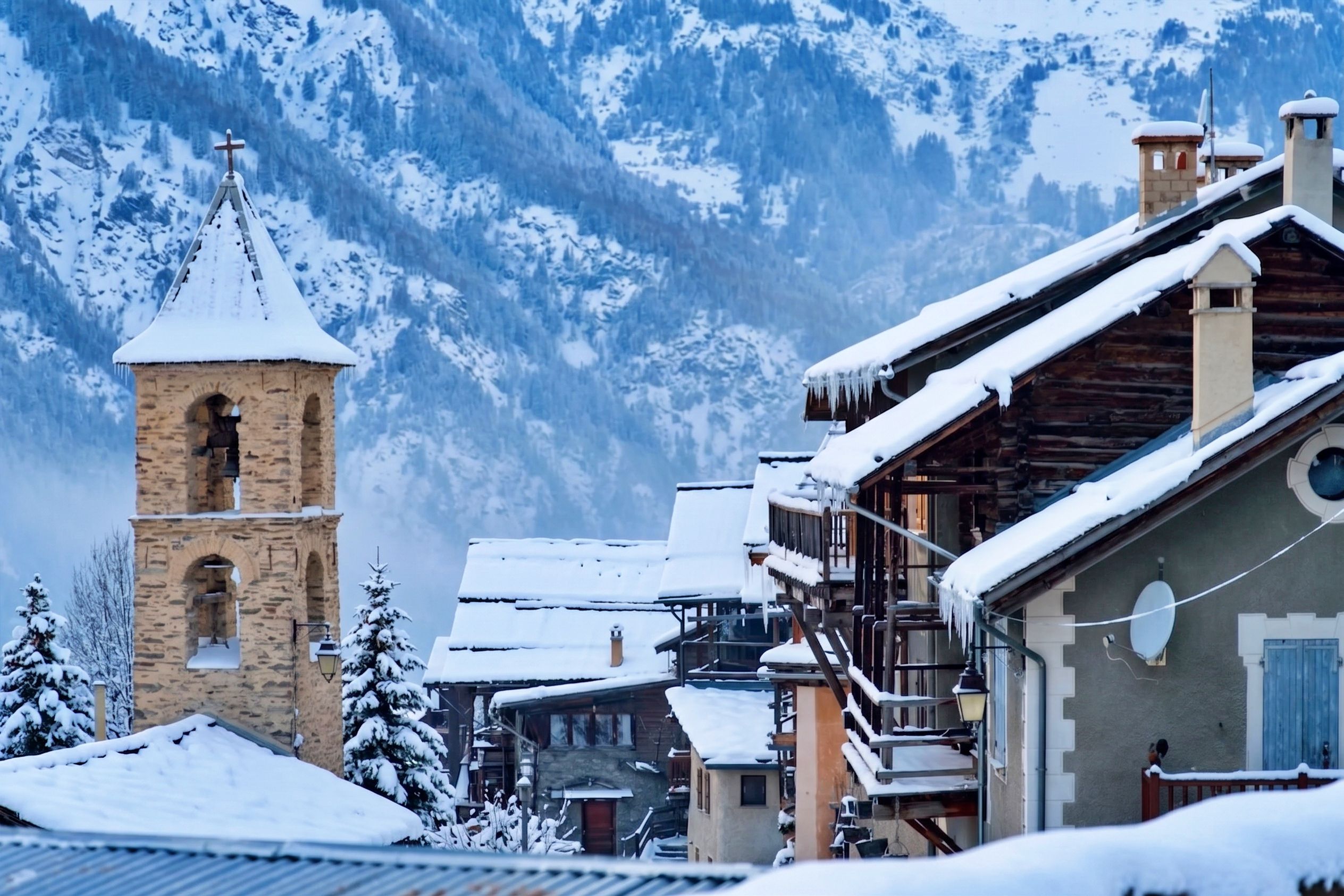 Een besneeuwd bergdorpslandschap met traditionele gebouwen, een klokkentoren en sneeuwtakken.