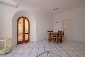 Ein Essbereich mit einem Holztisch und Stühlen, einem weiß gefliesten Boden, einer weißen Tür und einem Fenster mit Rundbogen.