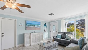 Ein Wohnzimmer mit Deckenventilator, einem Strand-Gemälde, einer Surfbrett-Dekoration, einem grauen Sofa mit Strandkissen und Blick auf Grünpflanzen durch große Fenster.