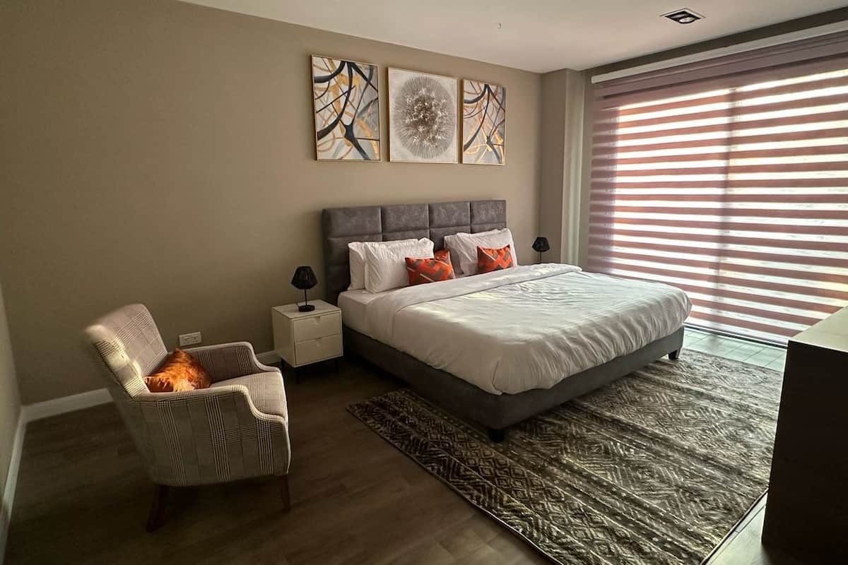 A bedroom with a bed, a chair, a nightstand, and a window with blinds.