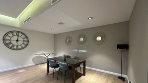A dining area with a wooden table and chairs, a wall clock, decorative wall art, recessed ceiling lighting, and a floor lamp. - Peravi Suites at Pearl In The City (Accra)