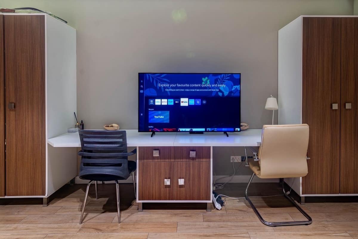 A modern room with a flat-screen TV mounted on the wall, a desk with two chairs, and wooden cabinets.