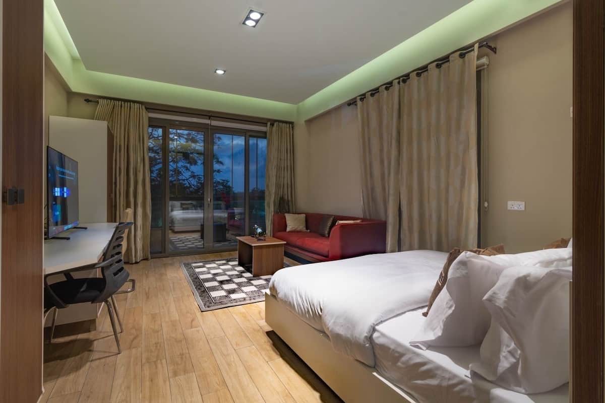 A modern hotel room with a bed, a desk, a red sofa, and a large window with curtains.