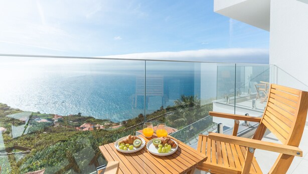 Een balkon met glazen hekwerk met uitzicht op de kust, met een houten tafel waarop croissants, fruit en een glas sinaasappelsap staan.
