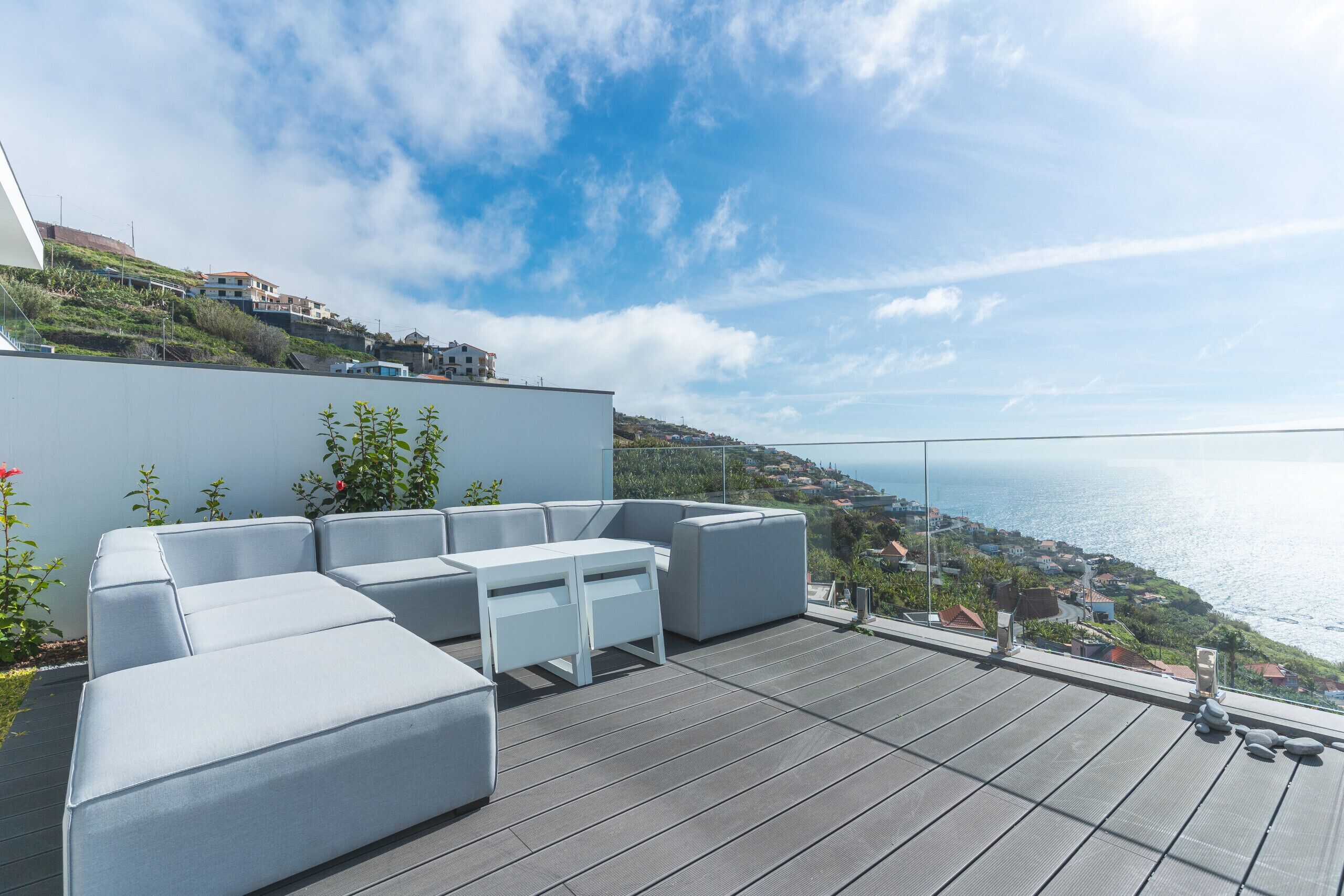 A rooftop terrace with a modern outdoor seating area, a clear view of the sea, and a hillside town in the background.