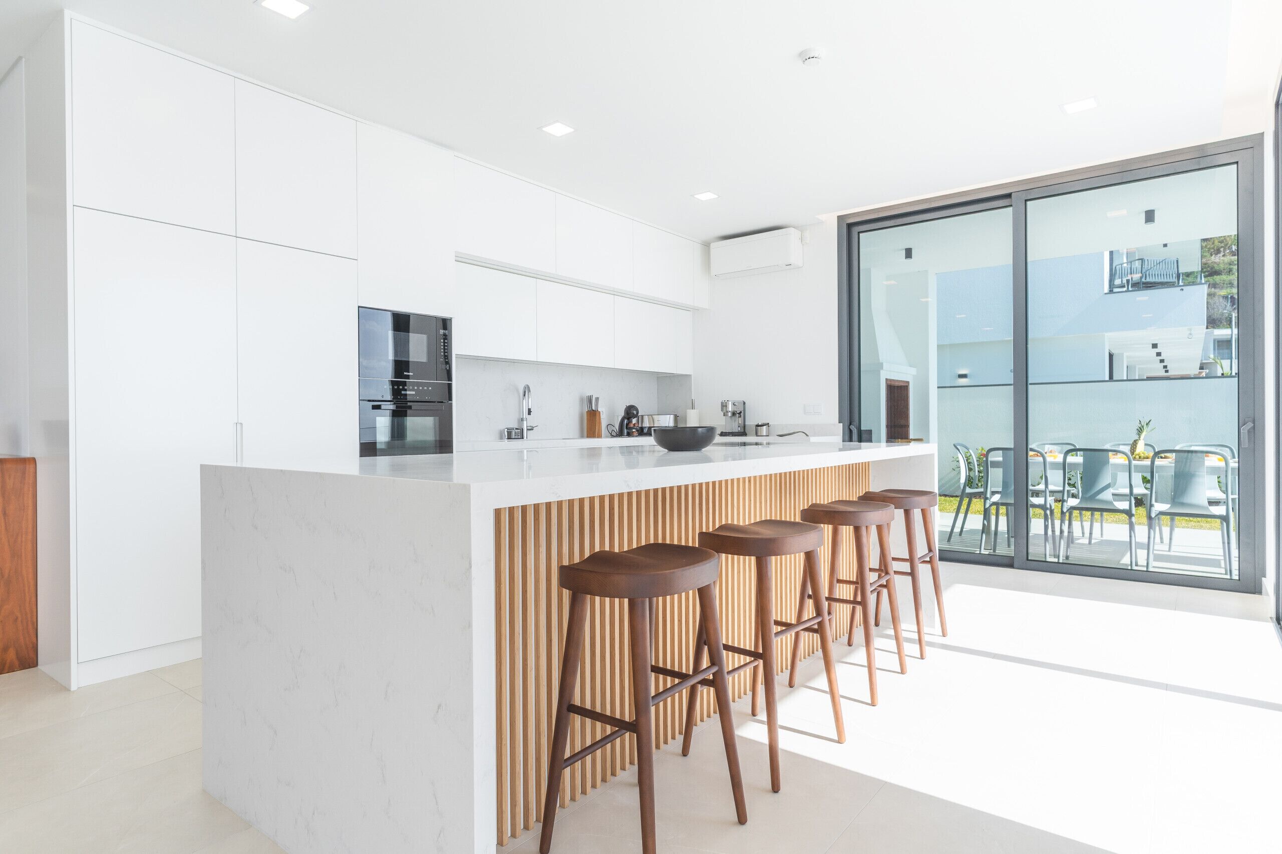 A modern kitchen with white cabinetry, a central island with stools, and a large sliding glass door leading to an outdoor area.