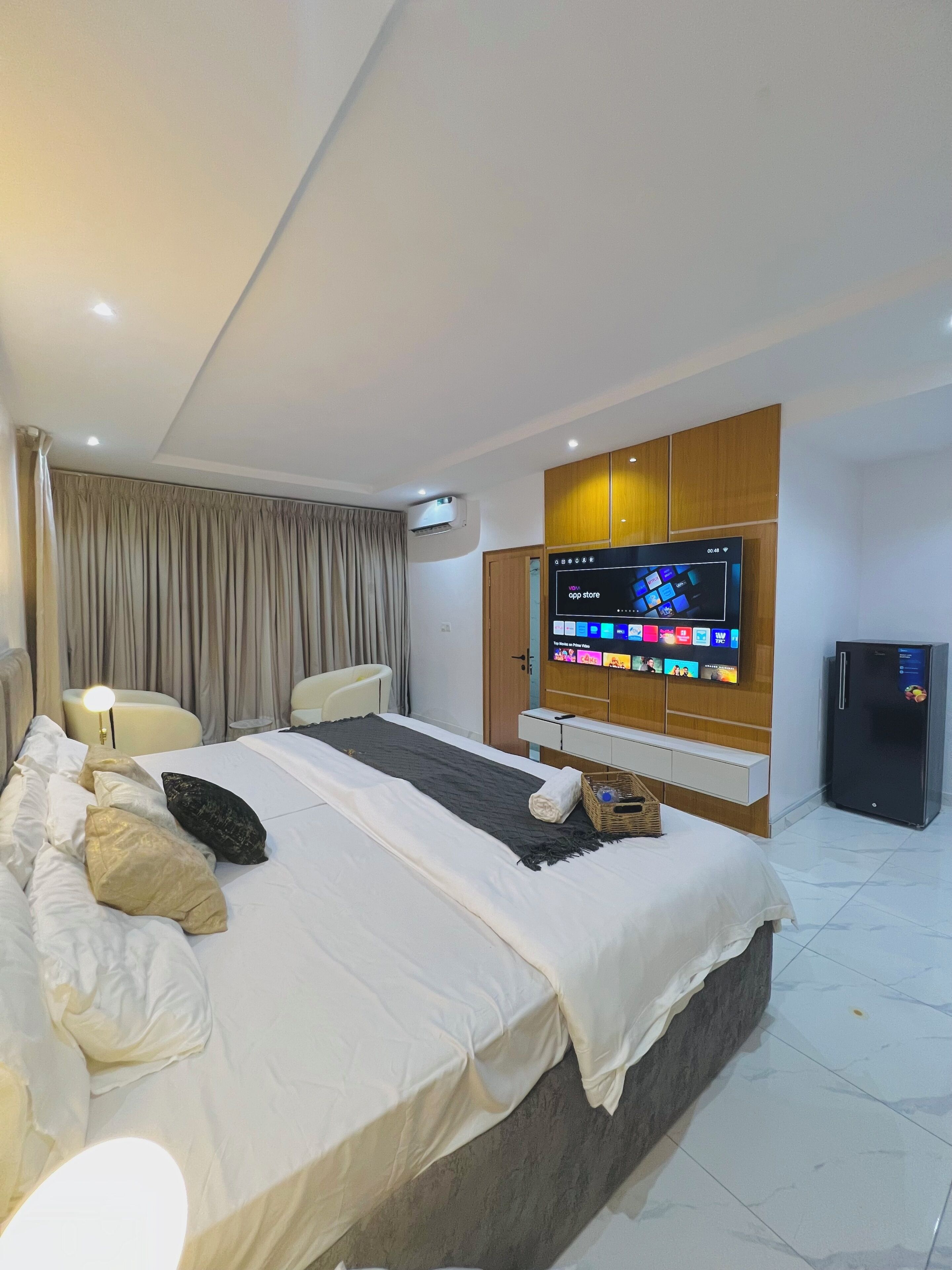 A modern hotel room with a large bed, a flat-screen TV mounted on the wall, a minibar, and a refrigerator.