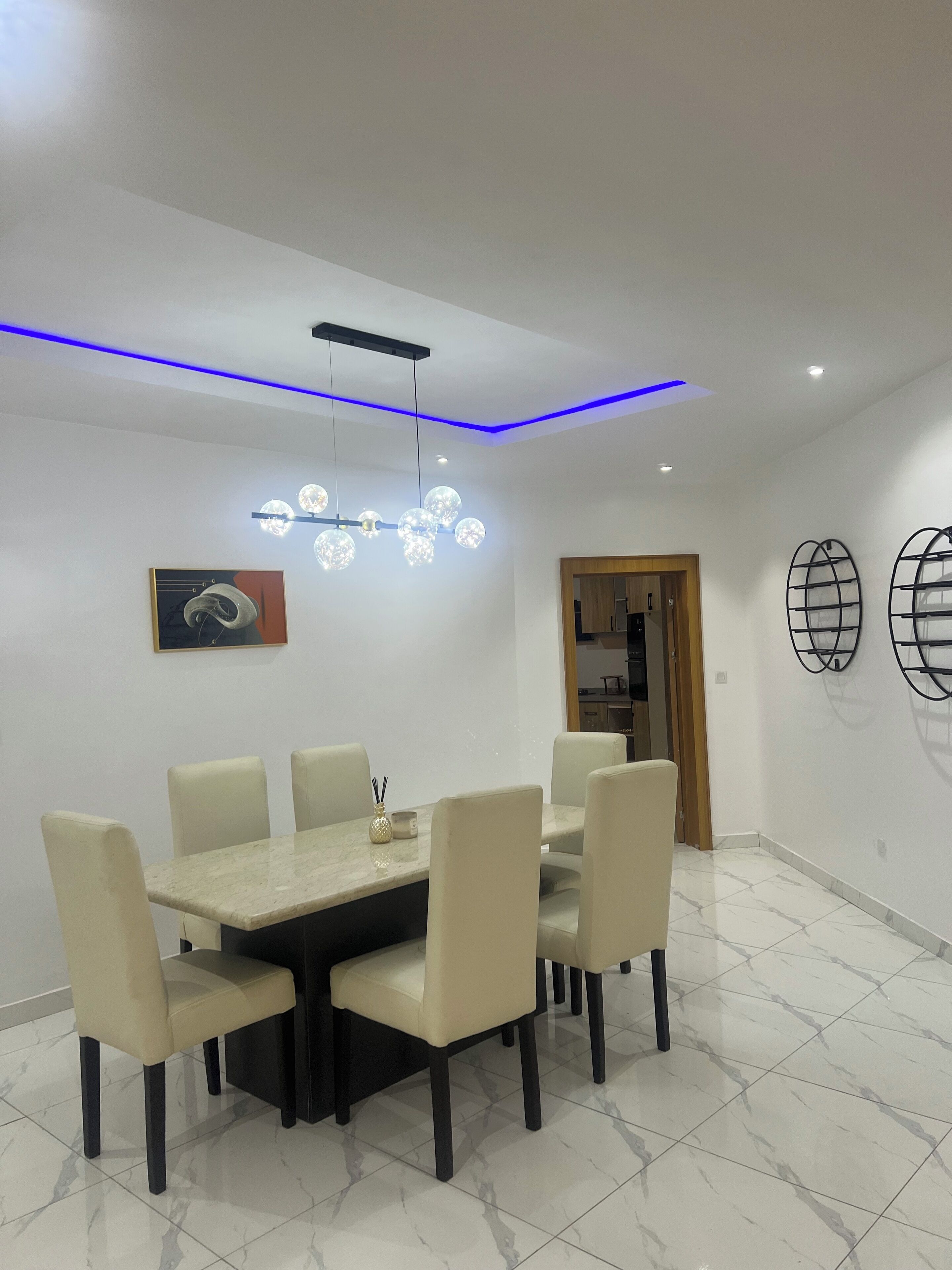 A dining area with a marble-tiled floor, a central table with six chairs, a modern chandelier, and wall-mounted shelves.