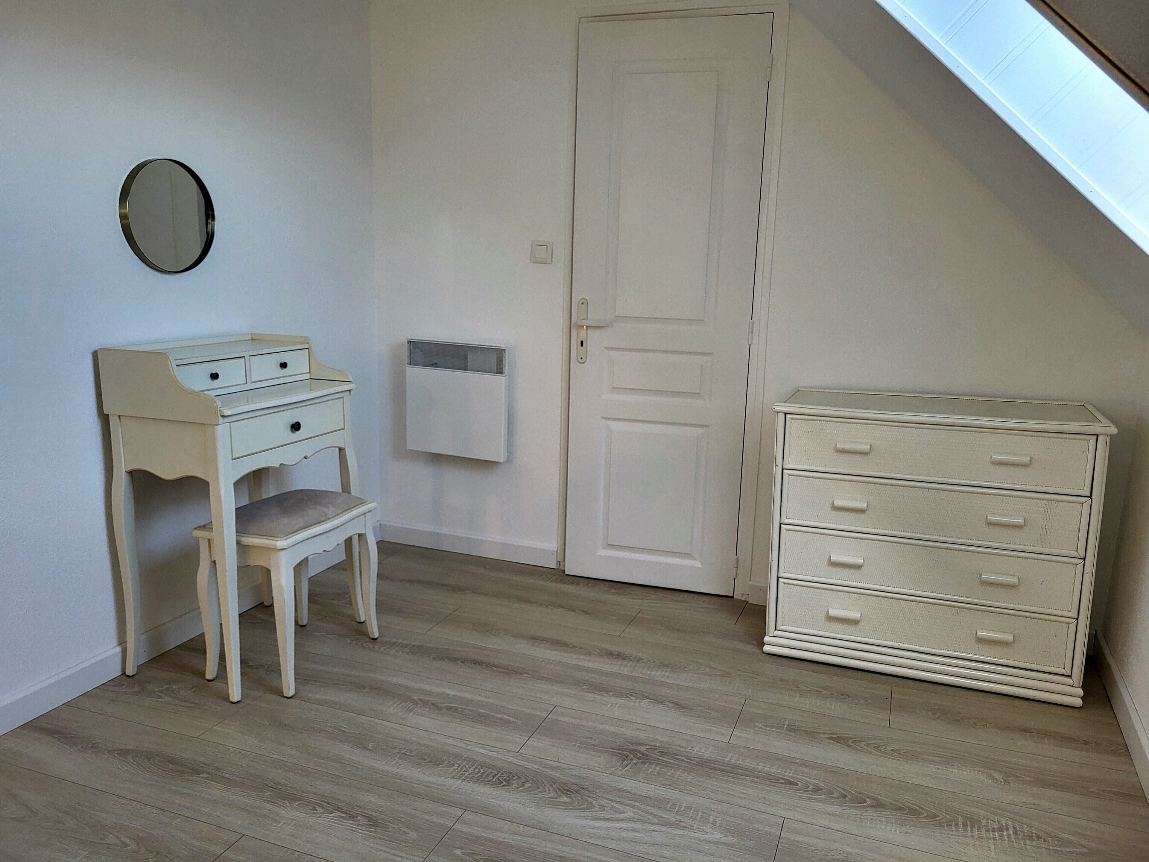 Ein Zimmer mit Schreibtisch, Stuhl, Kommode, einer Tür und einem kleinen Spiegel an der Wand.