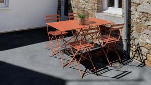 Ein Gebäude aus Stein mit einem Ziegeldach, einer weißen Außenwand und einer kleinen Terrasse mit Tisch und Stühlen.