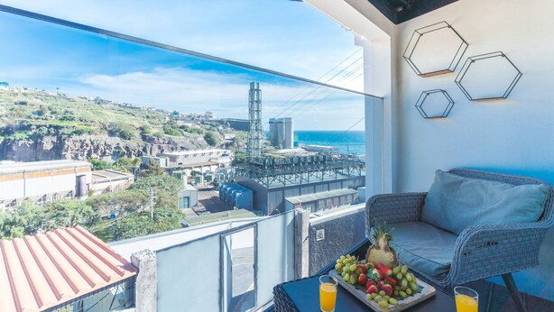 A balcony with a view of a coastal cityscape, including a bridge and industrial buildings. There is a wicker chair and a table with fruit and a glass of orange juice. - A Casinha by Atlantic Holiday (Câmara de Lobos)
