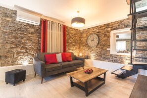 Ein modernes Wohnzimmer mit Steinwand, einem grauen Sofa mit roten Kissen, einem Holztisch und einem schwarzen Hocker.