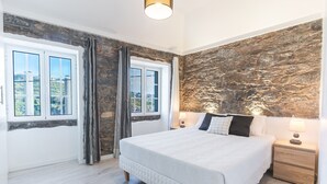 A bedroom with a stone wall, a large bed, two bedside tables with lamps, and a view through a window. - A Casinha by Atlantic Holiday (Câmara de Lobos)