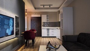 Un soggiorno moderno con un divano scuro, un tavolino da caffè con un vaso decorativo e una poltrona rossa. In evidenza, un televisore a parete mostra un'immagine. Sullo sfondo si intravede una cucina con mobili bianchi e uno schienale piastrellato.