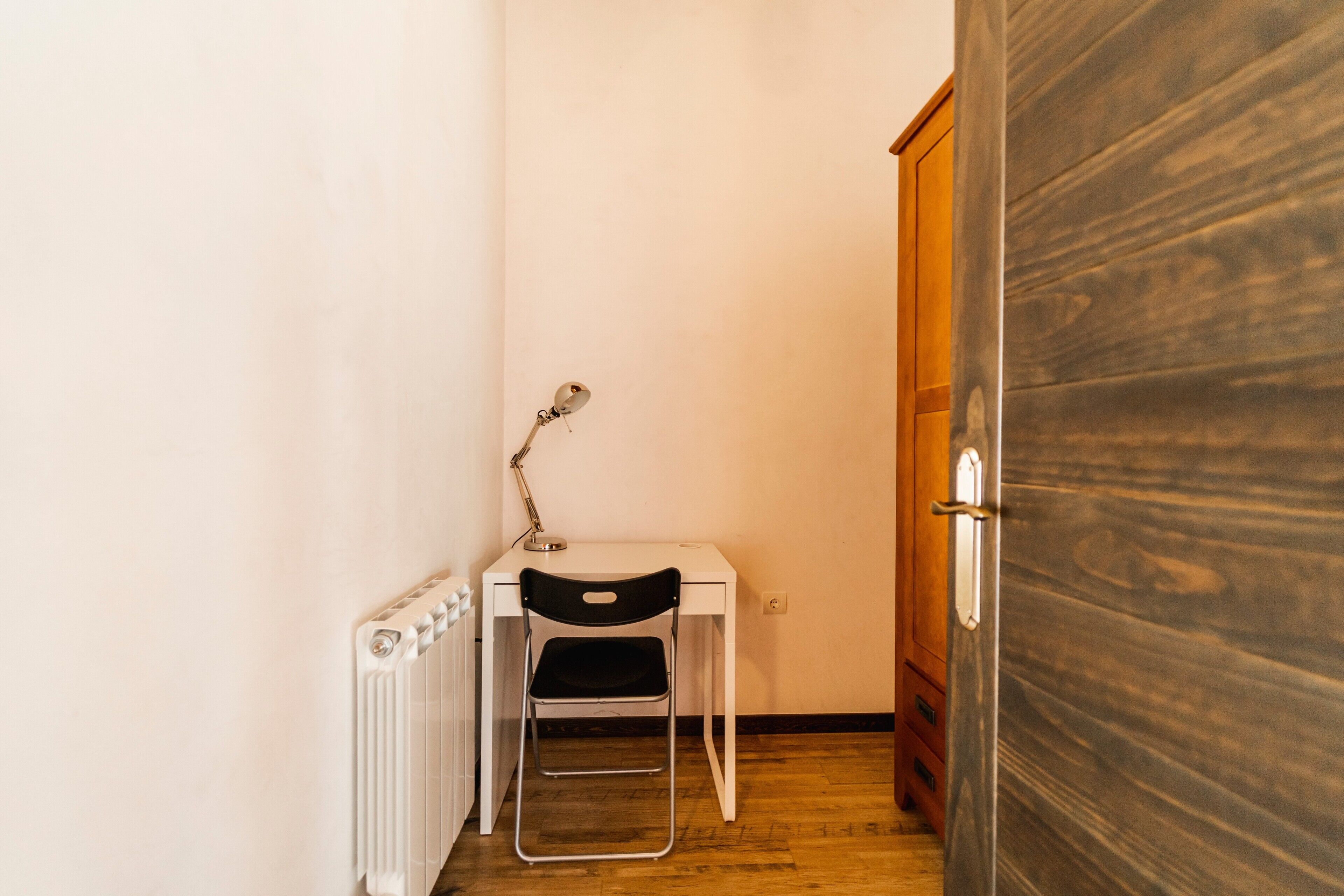 Um quarto pequeno com um radiador branco, uma cadeira preta, uma escrivaninha com uma lâmpada e uma porta de madeira.