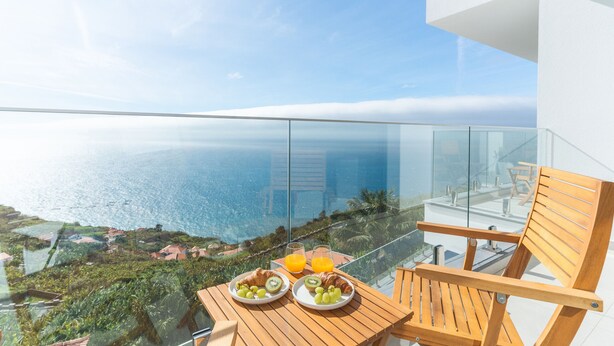 Ein Balkon mit Glasgeländer und Blick auf die Küste, mit einem Holztisch, der mit Croissants, Obst und einem Glas Orangensaft gedeckt ist.