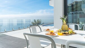 Ein Frühstückstisch mit Croissants, Ananas, Trauben und Orangensaft auf einem Balkon mit Blick aufs Meer.