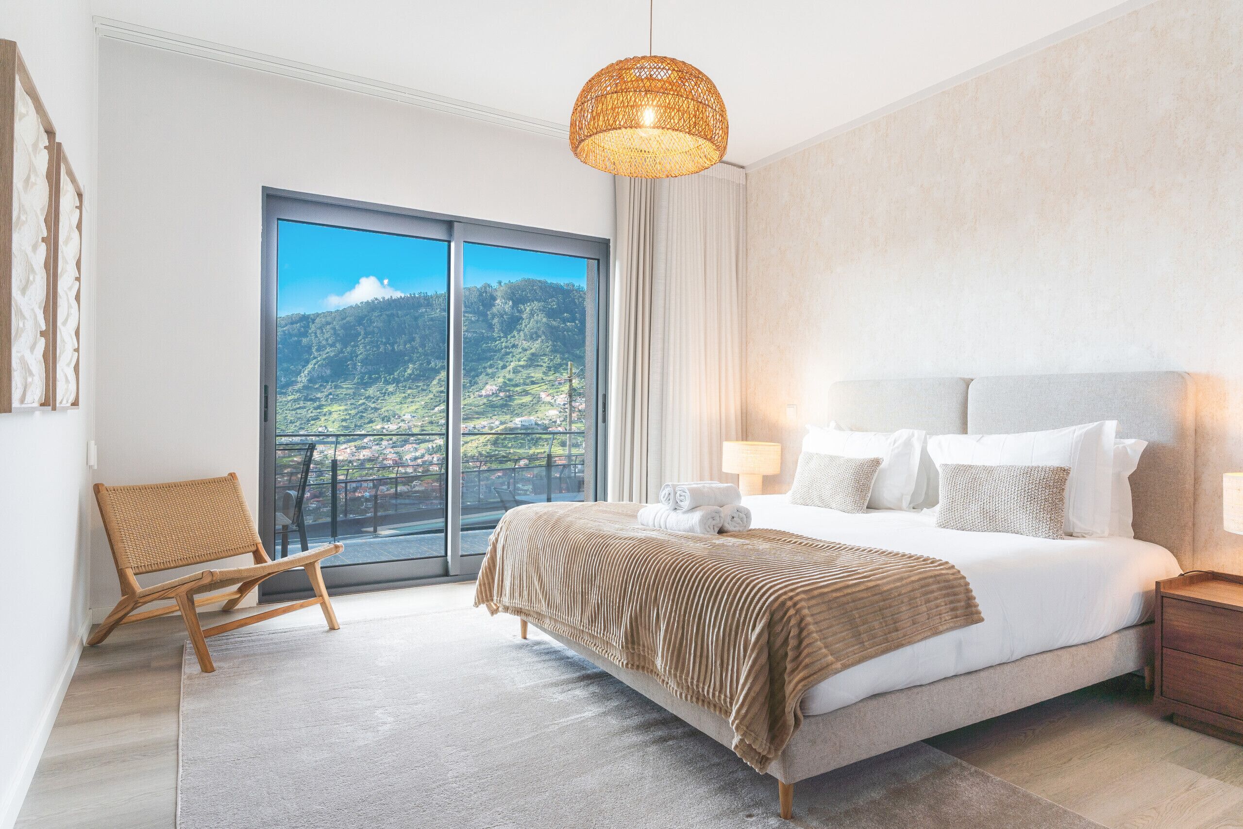 A modern bedroom with a large bed, a wicker chair, a balcony with a view of a mountain, and a hanging light fixture.