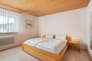 A bedroom with a wooden bed, a bedside table with a lamp, a radiator, and a window with sheer curtains.