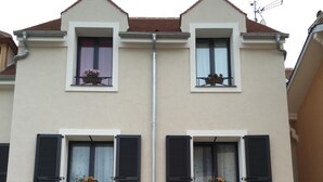 Ein zweistöckiges Gebäude mit einer symmetrischen Fassade, das Balkone mit Blumenkästen, eine Garage und eine auf dem Dach sichtbare Antenne aufweist.