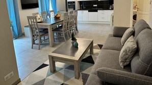 Ein modernes Wohnzimmer mit einer Couch, einem Esstisch mit Stühlen, einem Fernseher und einer Küche im Hintergrund.