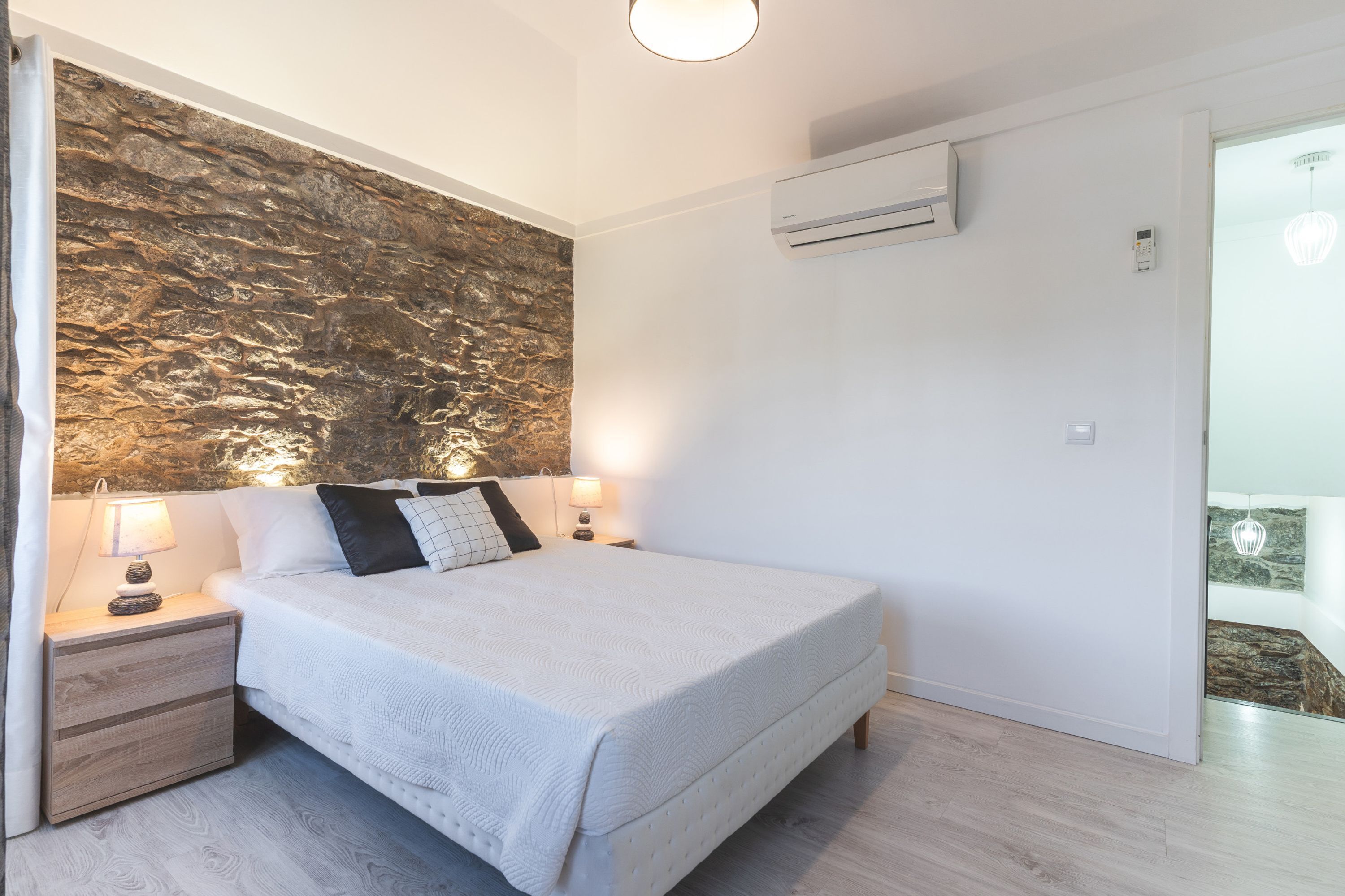 Un dormitorio con pared de piedra, una cama con ropa de cama blanca, dos lámparas de noche y un equipo de aire acondicionado en la pared.